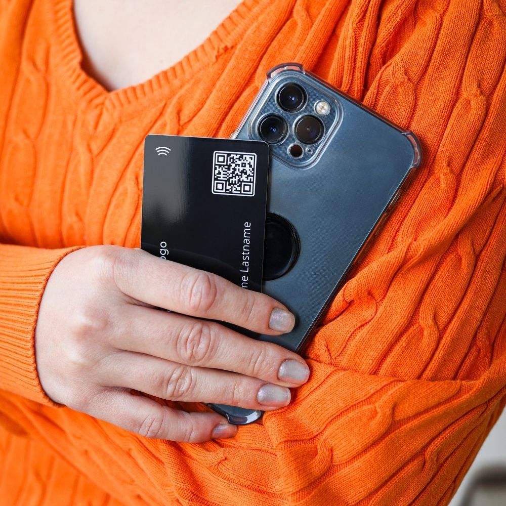 A woman wearing a organge sweater holds a mobile phone with a black nfc-sticker and a black NFC business card in her hand.