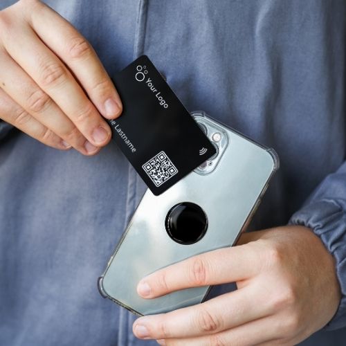 A woman wearing a blue blouse jacket holds a mobile phone with a black nfc-sticker a black the NFC business card in her hand.