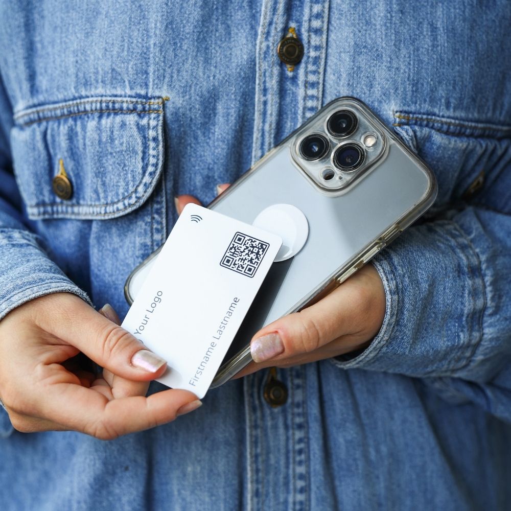 A woman wearing a blue denim jacket holds a mobile phone with a white nfc-sticker and a white NFC business card in her hand.