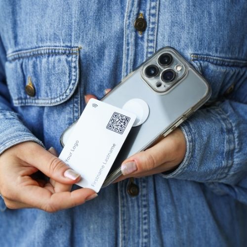 A woman wearing a blue denim jacket holds a mobile phone with a white nfc-sticker and a white NFC business card in her hand.