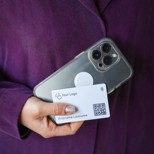 A woman wearing a purple sweater holds a mobile phone with a white nfc-sticker and a white NFC business card in her hand.